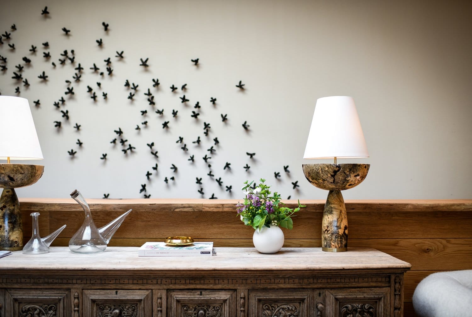 Elegant wooden sideboard with decorative lamps and a vase of flowers, wall adorned with artistic black bird sculptures.