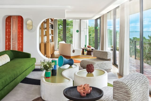 Modern living room with green sofa, circular tables, striped chairs, large windows, and colorful carpets.