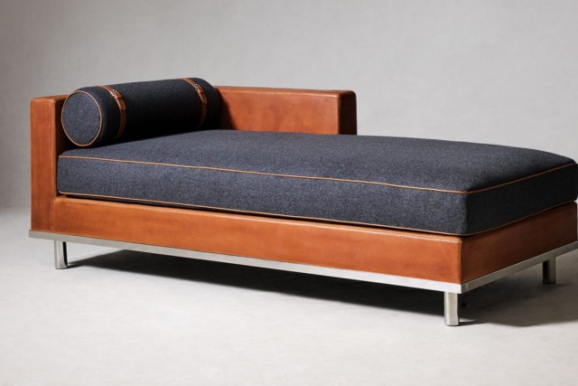 Modern chaise lounge with brown leather frame, dark fabric cushion, and cylindrical pillow on a metal base against a gray background.