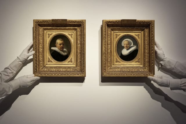 Gloved hands holding two framed oval portraits of older individuals with ruffled collars against a plain background.