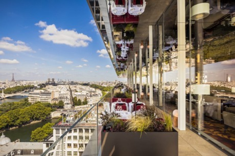 View of Paris skyline from rooftop restaurant with glass windows and greenery on terrace under a clear blue sky.
