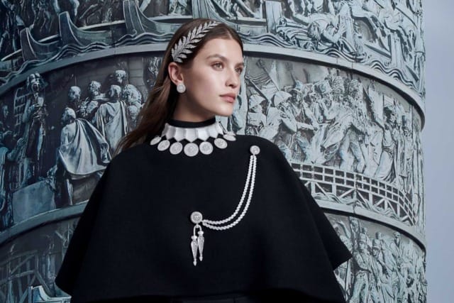 Model in elegant black outfit with silver jewelry, standing in front of a detailed, artistic relief backdrop.