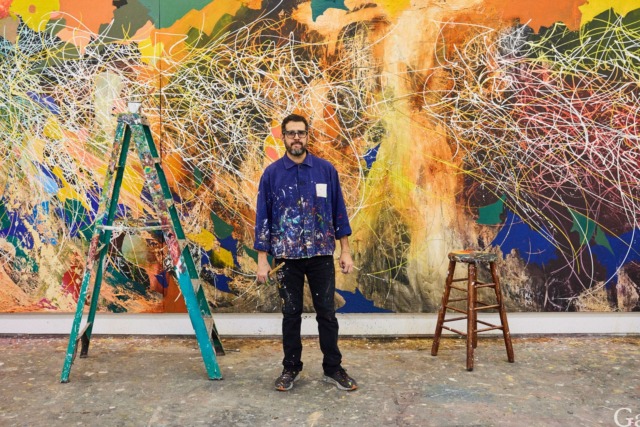 Artist standing in front of a large, colorful abstract painting with a green ladder and wooden stool to the side.