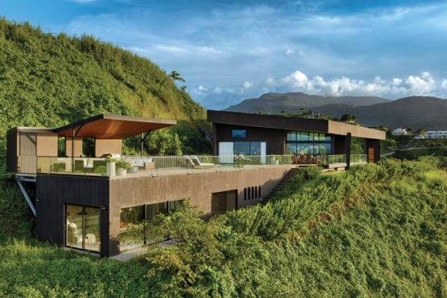 Modern hillside home with large glass windows, surrounded by lush greenery and mountains under a blue sky.