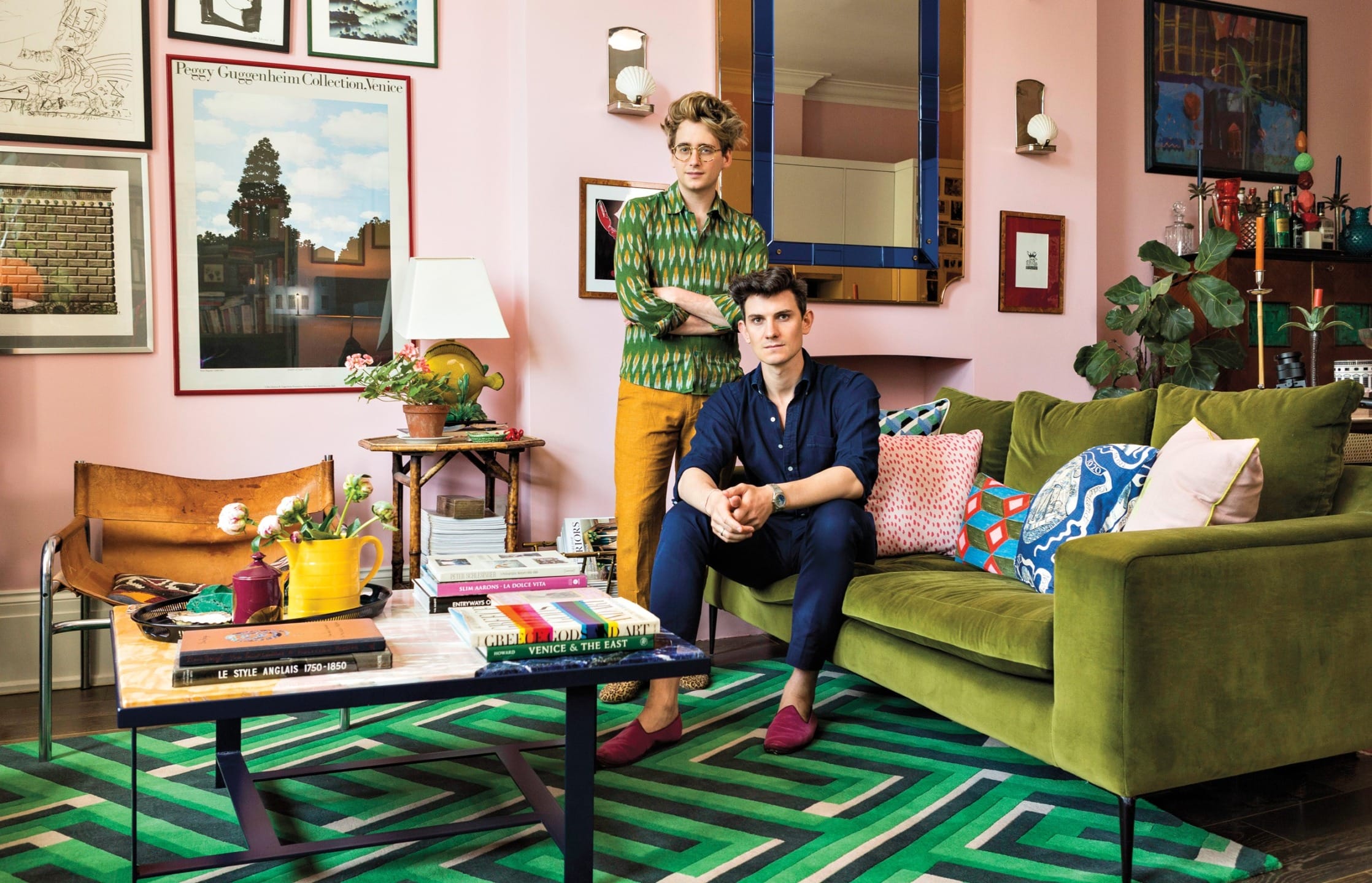 Two people in a colorful living room with vibrant decor, green velvet sofa, patterned rug, and framed artwork on the walls.