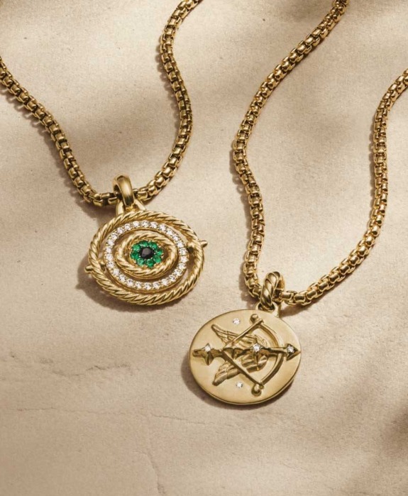 Two gold necklaces on a beige background, one with a green gem and intricate design, the other round with a symbolic engraving.