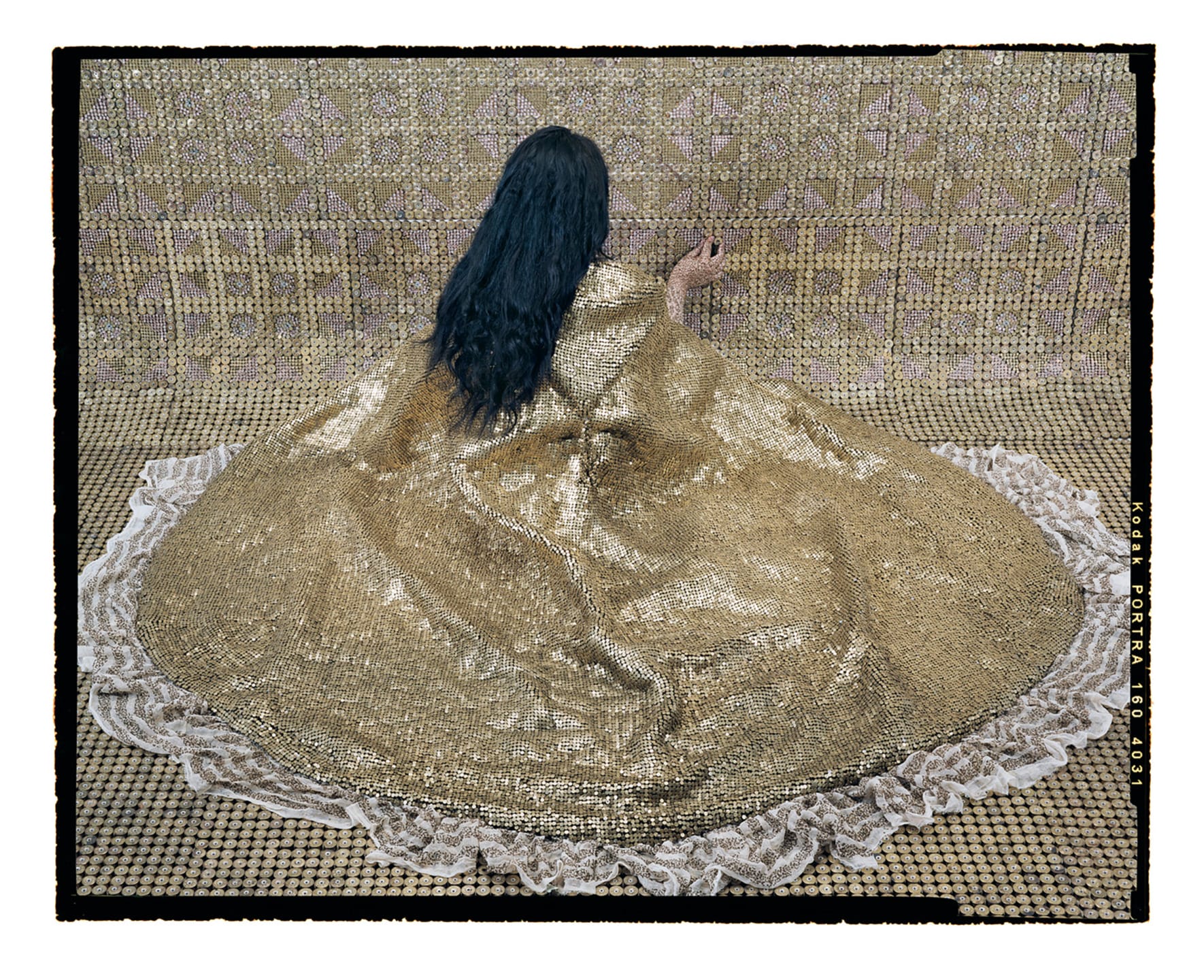 Woman with long dark hair in a golden intricate dress sits on a matching ornate background, facing away.