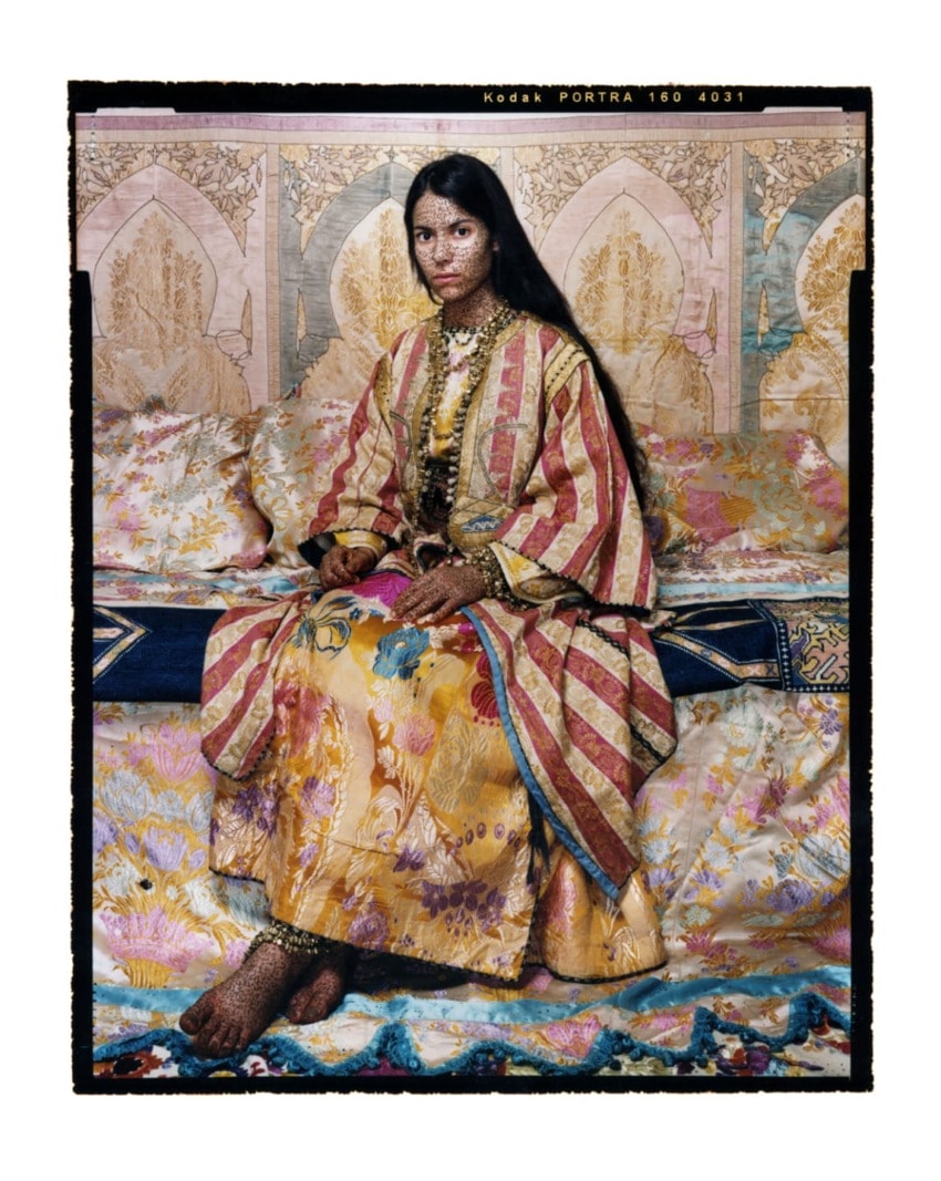 Person in ornate, colorful traditional clothing seated against a decorative background.