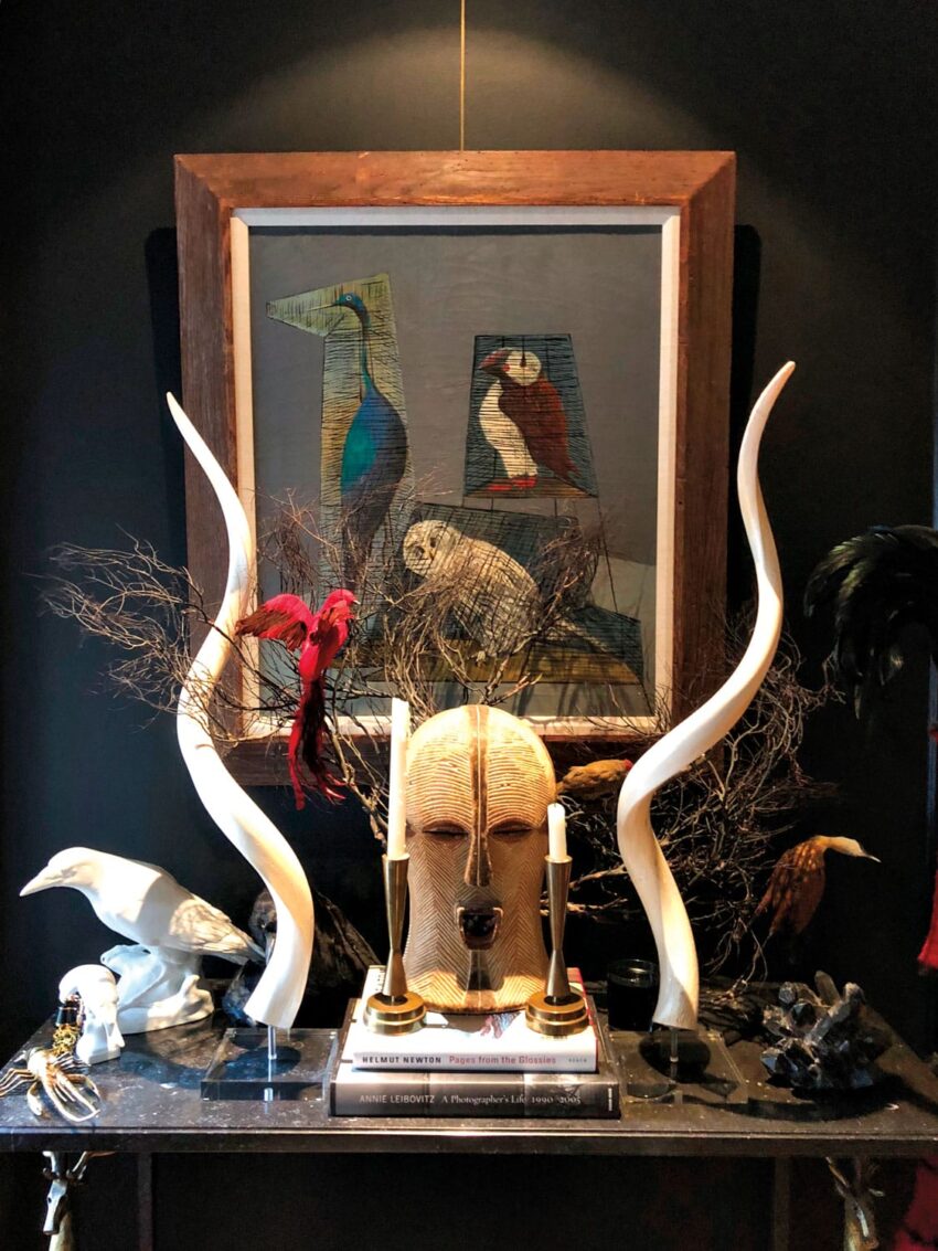 Decorative mask with horns, framed bird artwork, red bird, and various sculptures on a dark wall shelf.