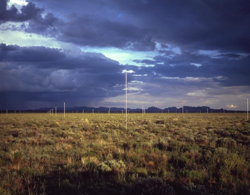 Wide open grassland under a dramatic cloudy sky with white poles scattered across the landscape and distant mountains.