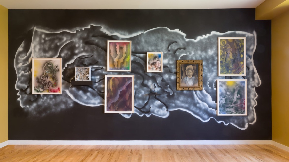 Wall art with multiple framed abstract paintings on a dark mural background in a bright room with wooden flooring.