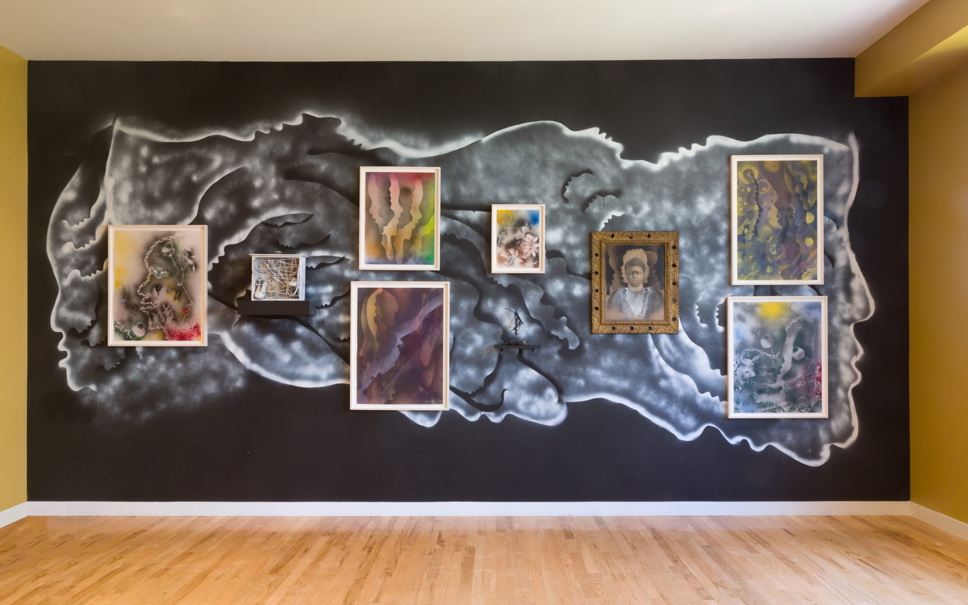 Wall art with multiple framed abstract paintings on a dark mural background in a bright room with wooden flooring.