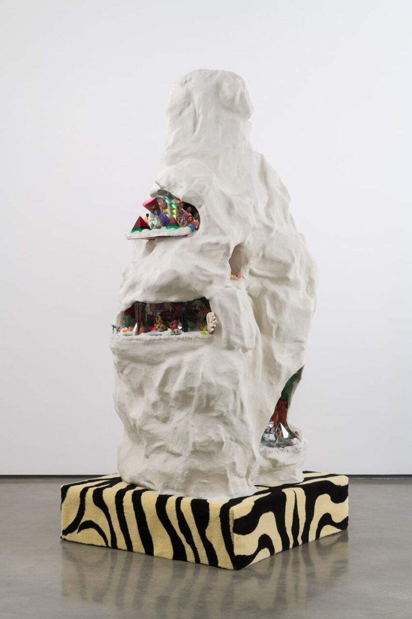 Large white sculpture with vibrant miniature scenes inside, displayed on a zebra-striped platform in a gallery setting.