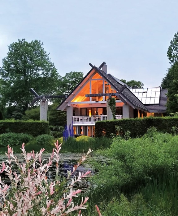 Modern house with large windows surrounded by lush greenery and a pond in the foreground.