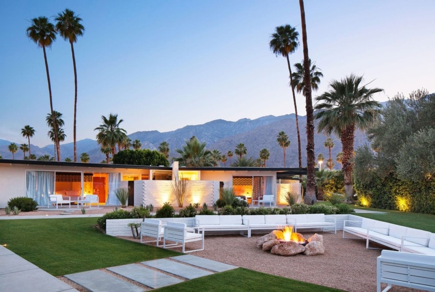 Mid-century modern house with palm trees, mountain view, and outdoor seating around a fire pit at dusk.