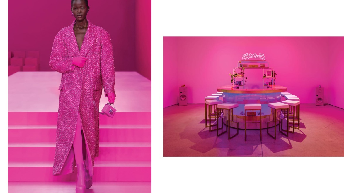 Model in a long pink coat on a runway, next to a pink-themed room with a circular table and stools.