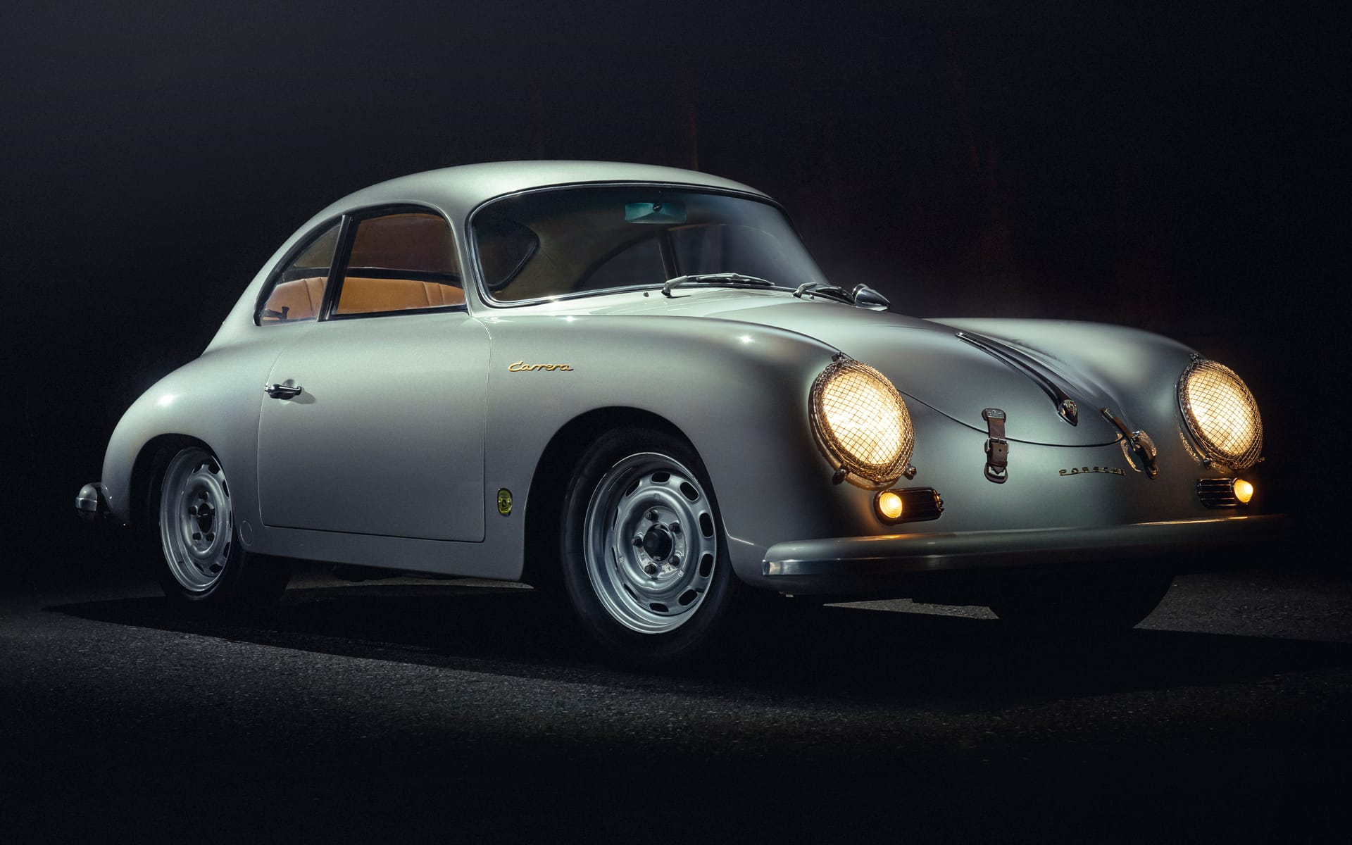 Vintage silver sports car with rounded design, illuminated headlights, and classic wheels parked on a dark road background.