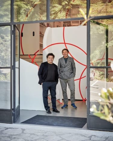 Two men standing in an open doorway with a modern interior featuring red geometric designs in the background.