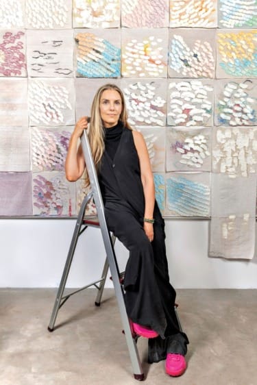 Person sitting on a ladder in front of a wall with abstract textile art, wearing a black dress and bright pink shoes.
