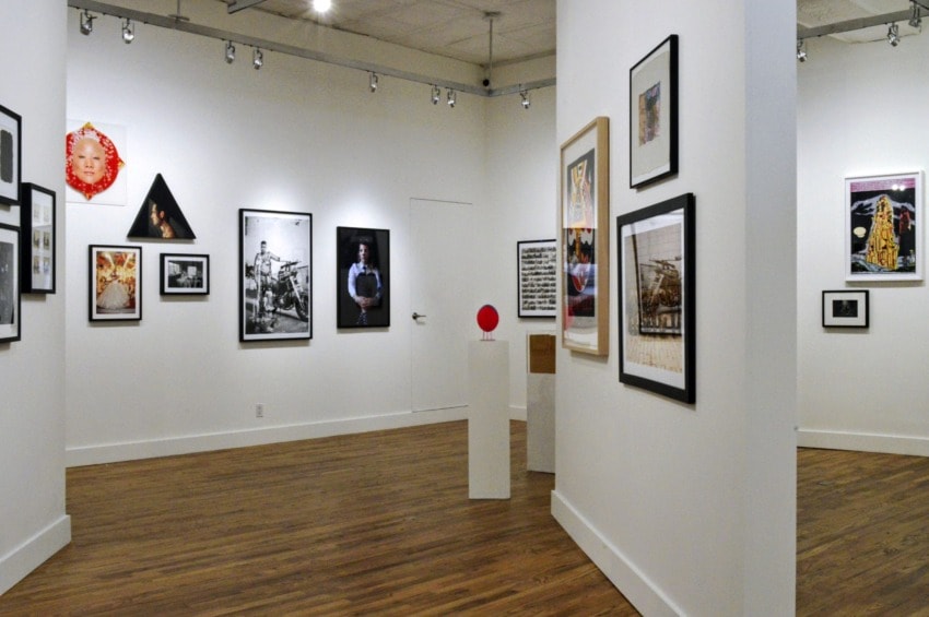 Art gallery interior with various framed artworks on white walls and a sculpture on a pedestal, wooden floor.