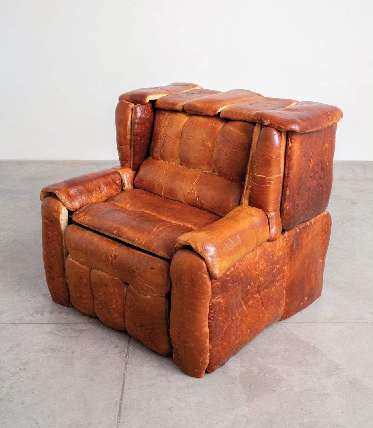 Armchair designed to look like it's made of bread, placed in a minimalist room with a concrete floor.