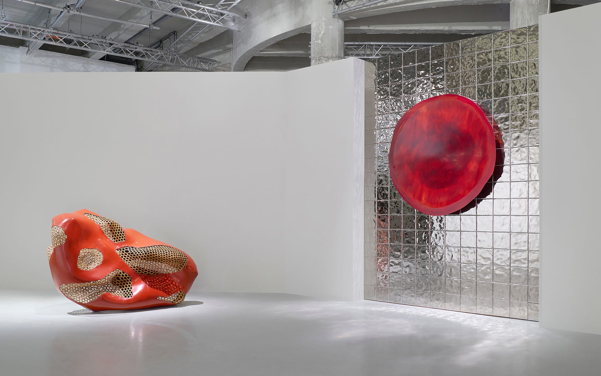Abstract art installation with a red sculpture and a circular red piece on a glass block wall in a modern gallery space.