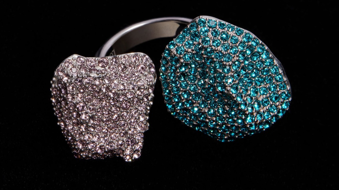 Silver ring with two textured stones, one covered in small clear gems and the other in sparkling turquoise gems, on a black background.