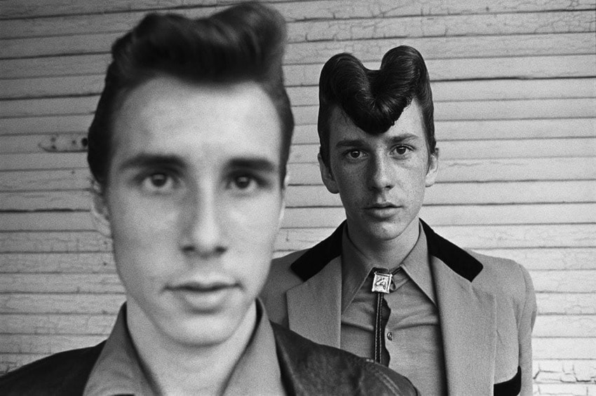 Two men with vintage hairstyles and attire stand in front of a textured wall, one in focus and the other blurred in the foreground.