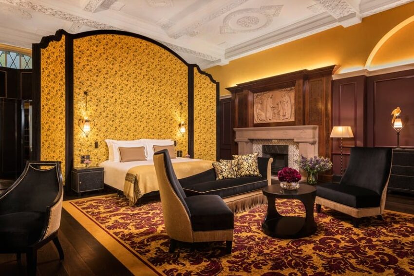 Luxurious hotel room with ornate decor, featuring a bed, elegant chairs, fireplace, and intricate ceiling design.