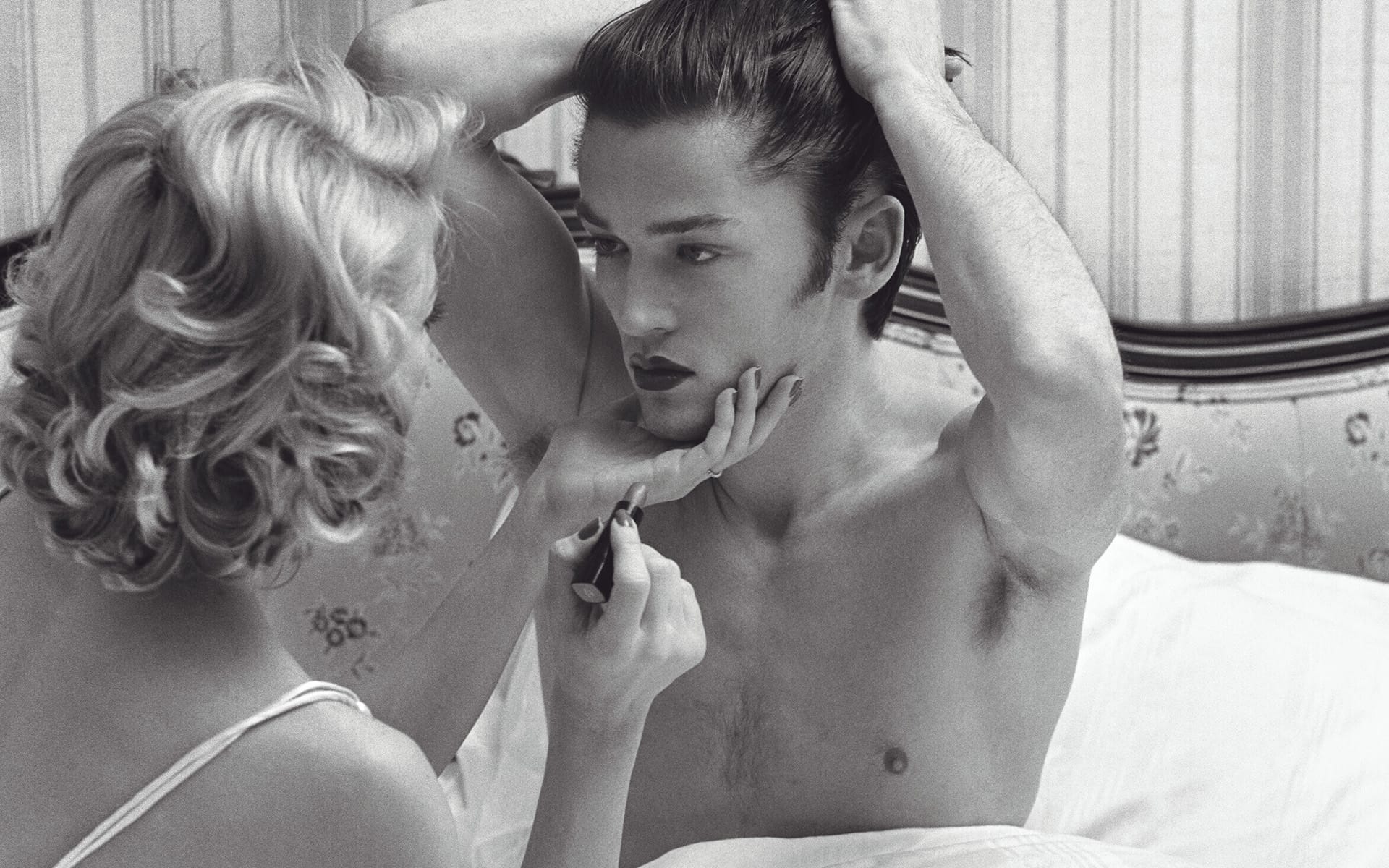 A man sitting on a bed getting his lipstick applied by a woman in a black and white setting.