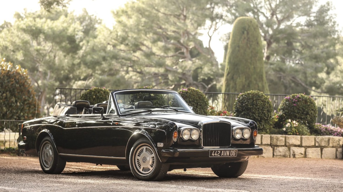 Luxury black convertible car parked in a scenic area with trees and flowers in the background.