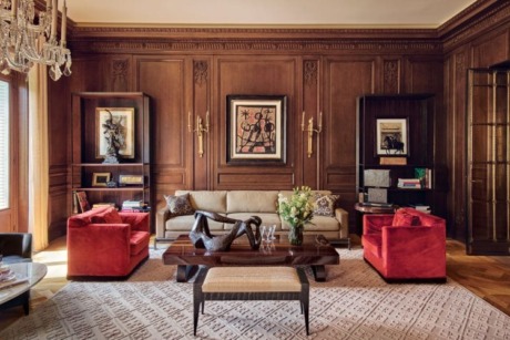 Elegant living room with wood-paneled walls, beige sofa, red armchairs, art pieces, and a chandelier.