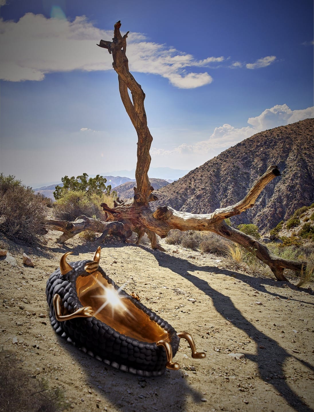 Artistic display of a sculpture resembling a curled human form under a twisted tree in a desert landscape with mountains.