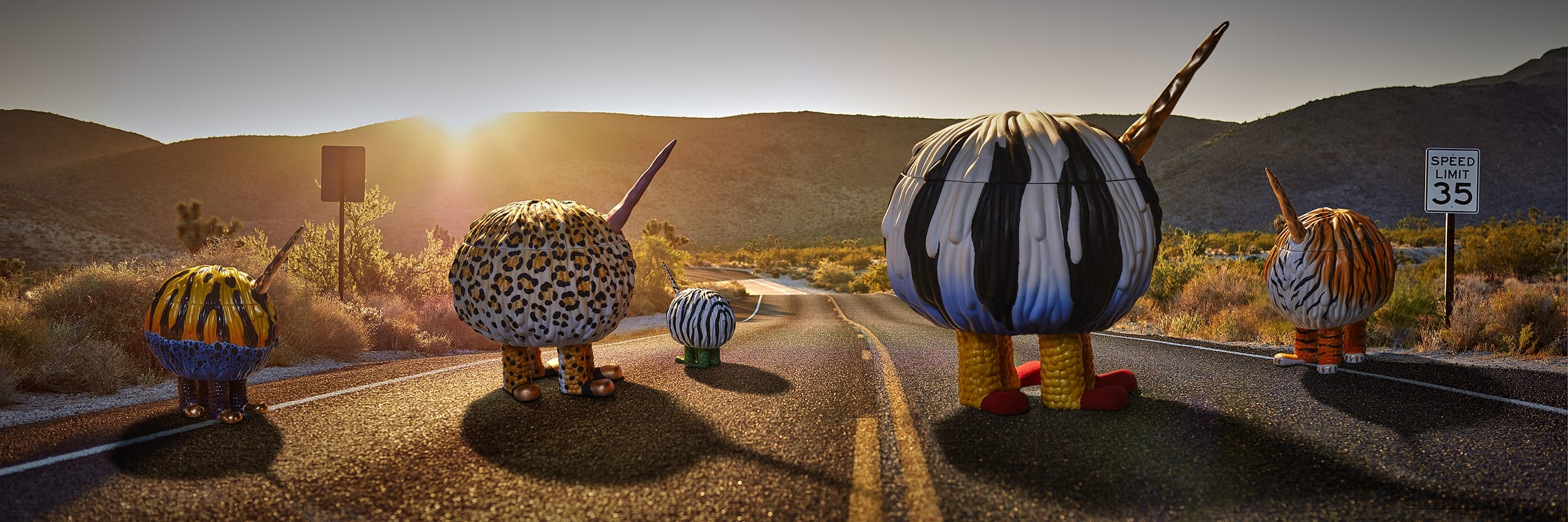 Colorful, round figures with striped patterns walk down a desert road at sunset, passing a speed limit sign.