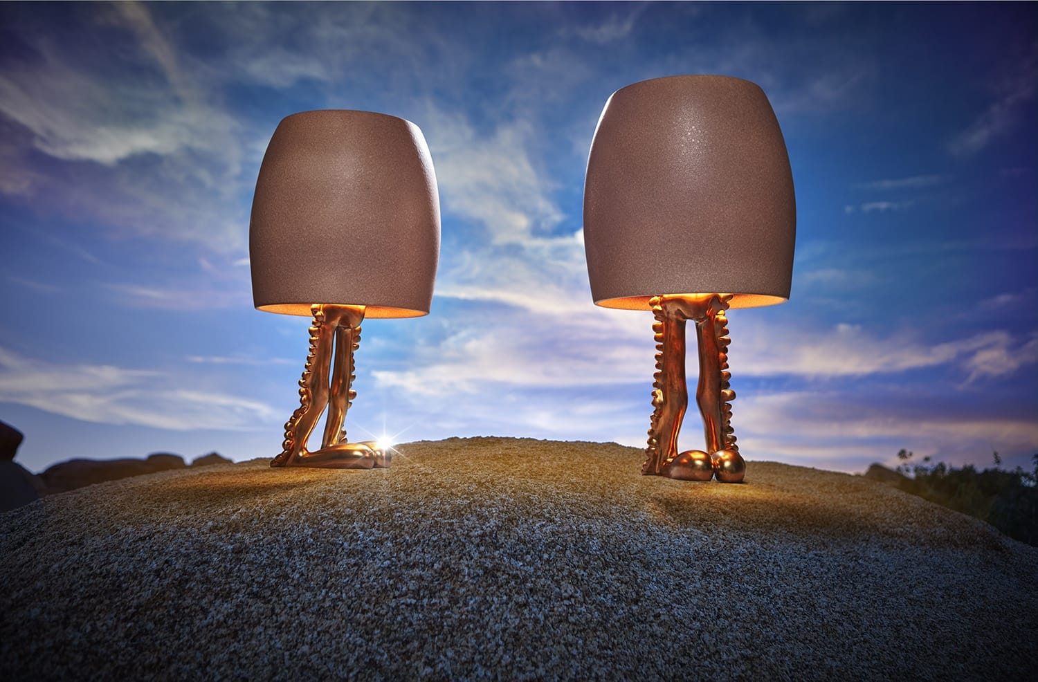 Two artistic lamps with stylized legs as bases, standing on a sandy surface against a vibrant blue sky at sunset.