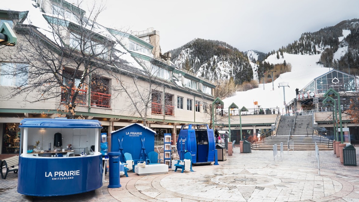 Outdoor shopping area in winter with snow-covered mountains, blue kiosks, and empty seating.