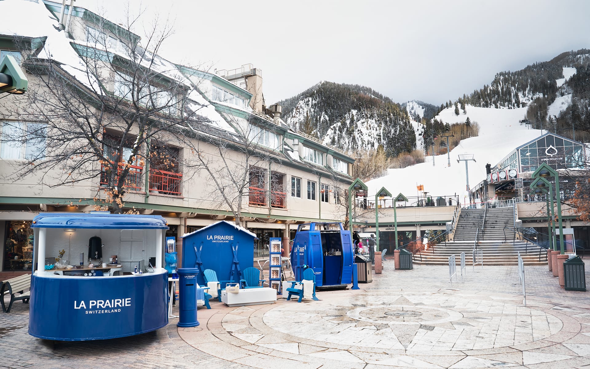 Outdoor shopping area in winter with snow-covered mountains, blue kiosks, and empty seating.