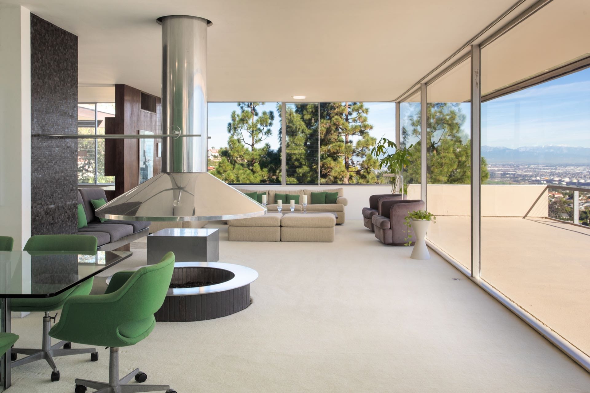 Modern living room with large windows, green chairs, and a central metallic fireplace overlooking a scenic outdoor view