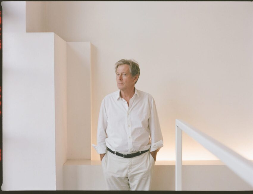 A person in a white shirt stands in a minimalist white room with geometric wall features, hands in pockets.
