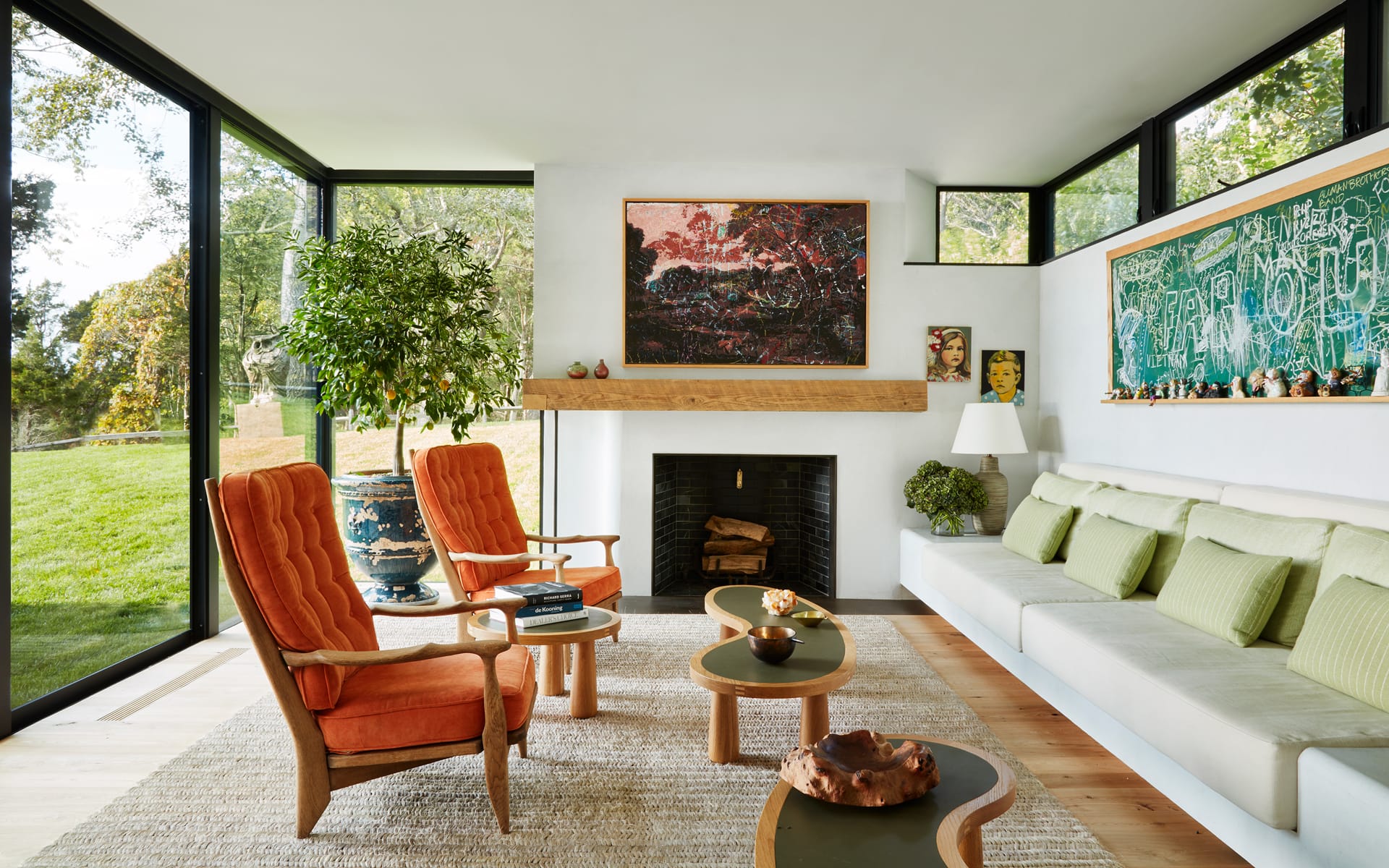 Modern living room with large windows, orange chairs, a fireplace, and greenery, featuring colorful artwork on the walls.