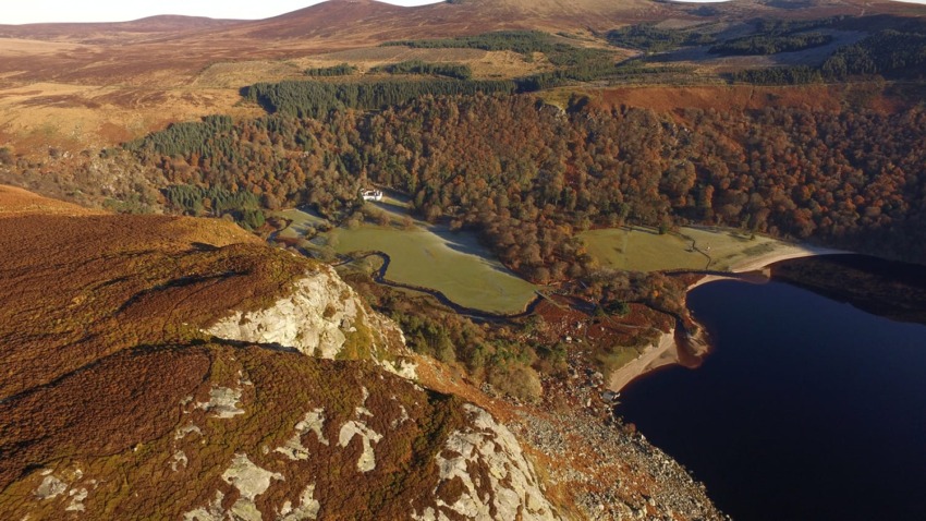 Aerial view of a scenic landscape with a lake, forest, and rocky cliffs under a clear sky in autumn.