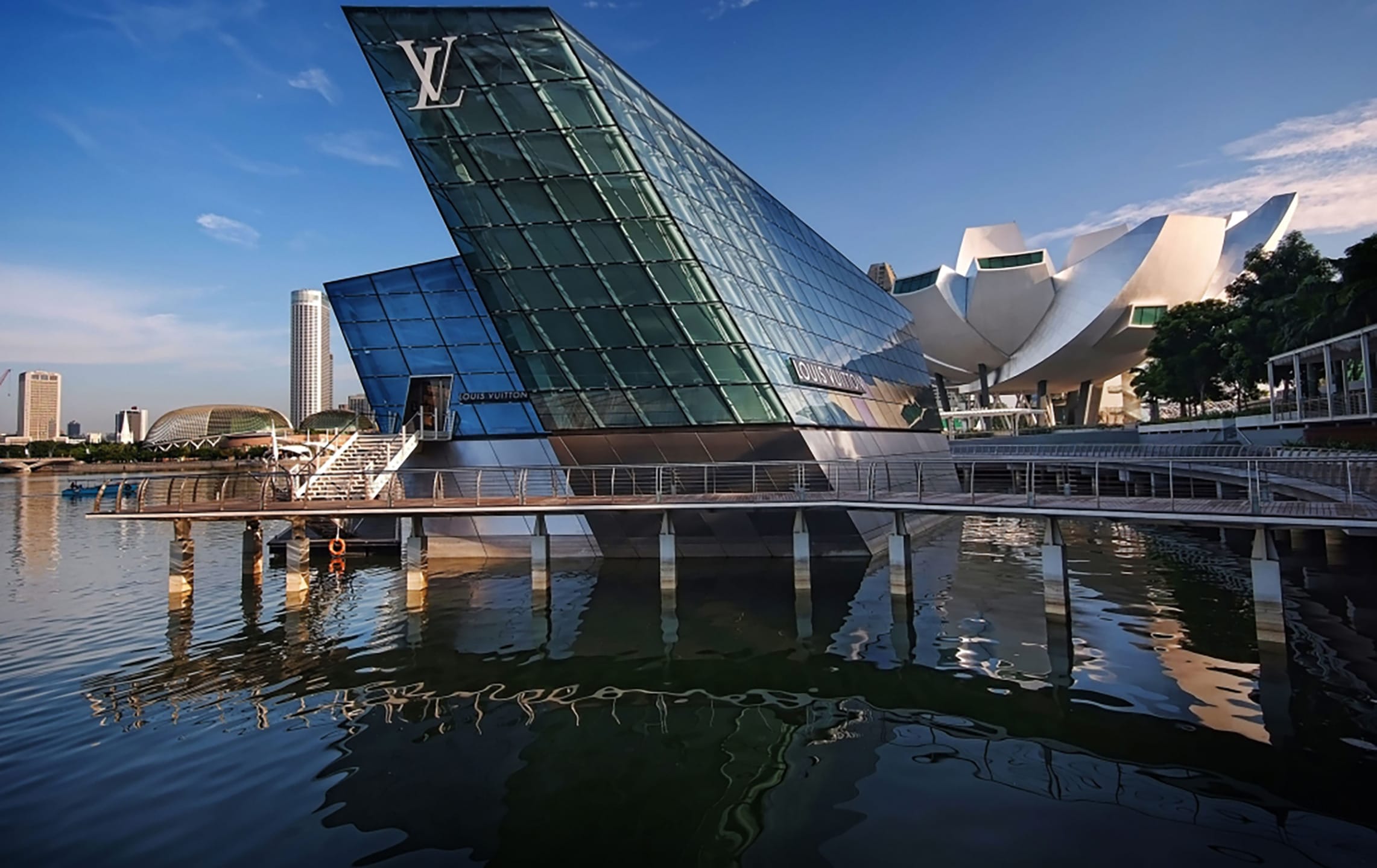 Modern glass building with geometric design over water in an urban setting, featuring reflections and a clear blue sky.