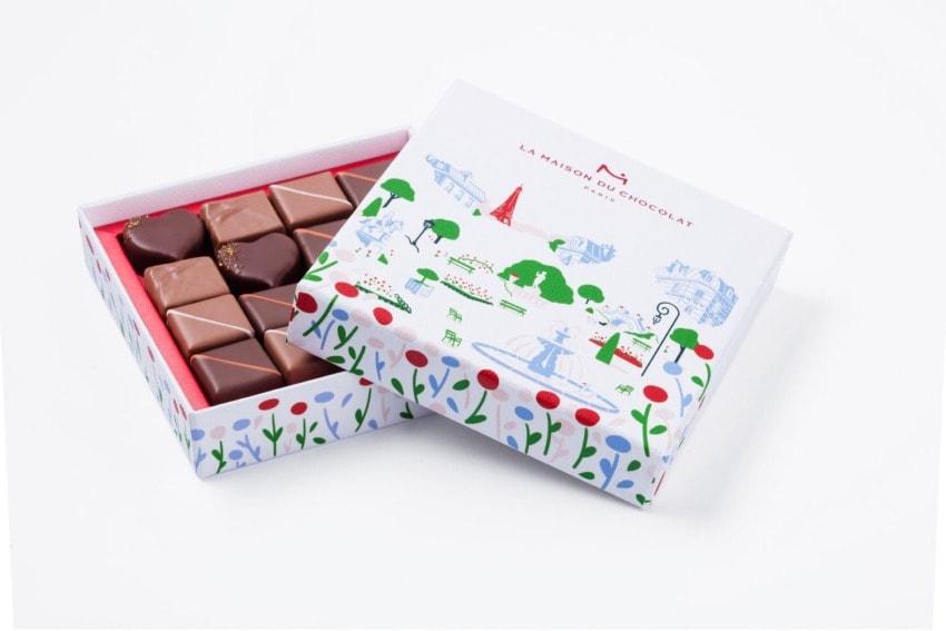 Box of assorted chocolates with a colorful, illustrated lid partially open to reveal heart-shaped and square chocolates.