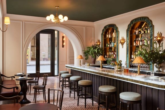 Cozy elegant bar interior with vintage decor, arched doorway, marble counter, lush plants, and warmly lit shelves.