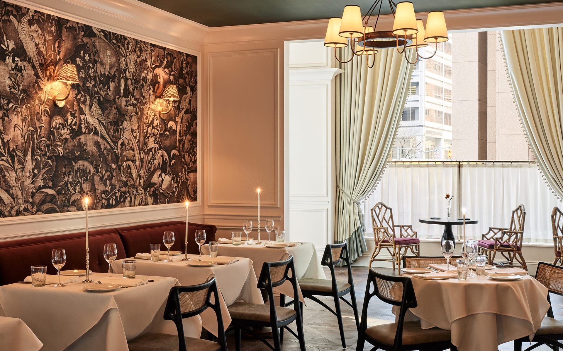 Elegant restaurant interior with neatly set tables, wall art, chandeliers, and large window draped with curtains.