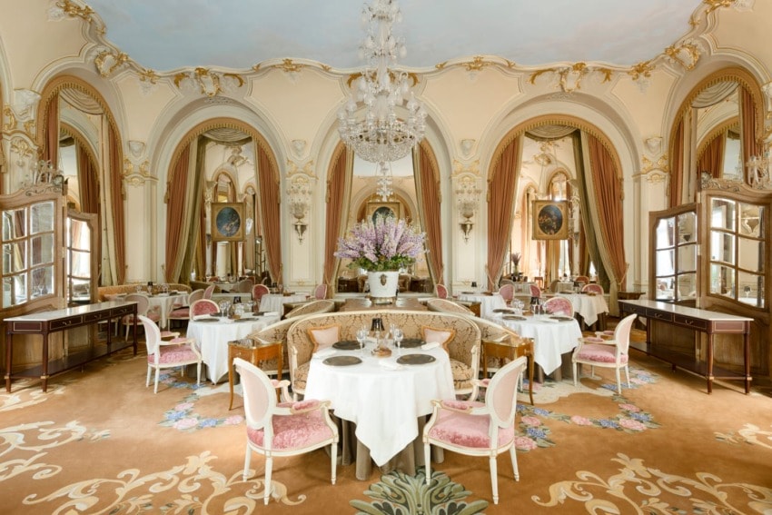 Elegant restaurant dining room with ornate décor, chandeliers, and pink velvet chairs arranged around white tablecloth-covered tables.