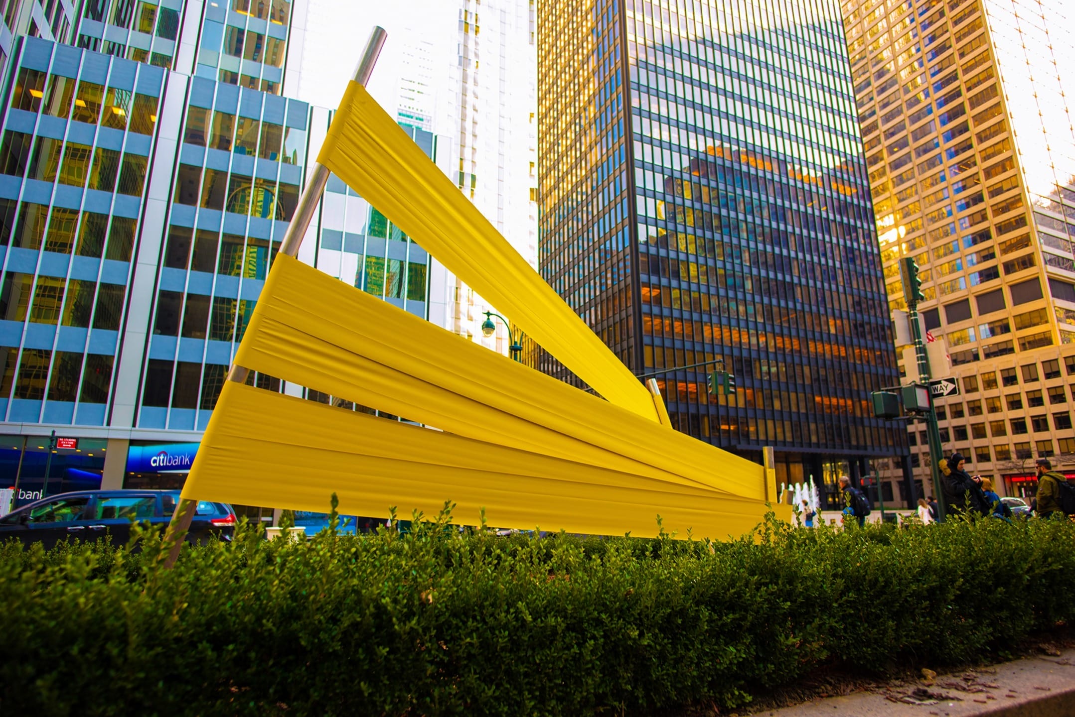 Yellow abstract sculpture with angled beams in a city plaza, surrounded by modern glass skyscrapers and greenery.