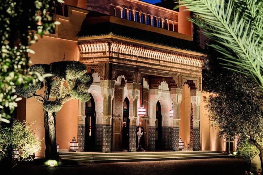 Ornate Moroccan building entrance illuminated at night, featuring detailed arches and a manicured tree in the foreground.