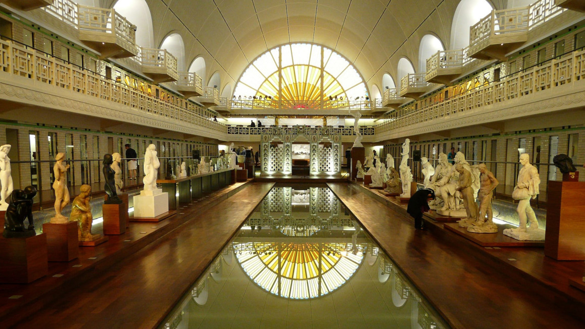 Art sculptures displayed around a pool with a large, arched window in the background at a museum exhibition hall.