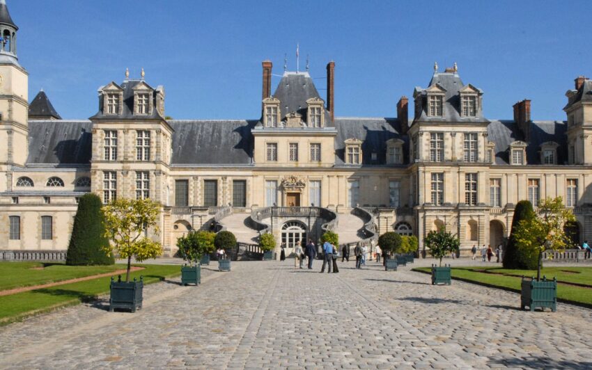 People walking in front of Château de Fontainebleau, with its historic architecture and symmetrical gardens on a sunny day.